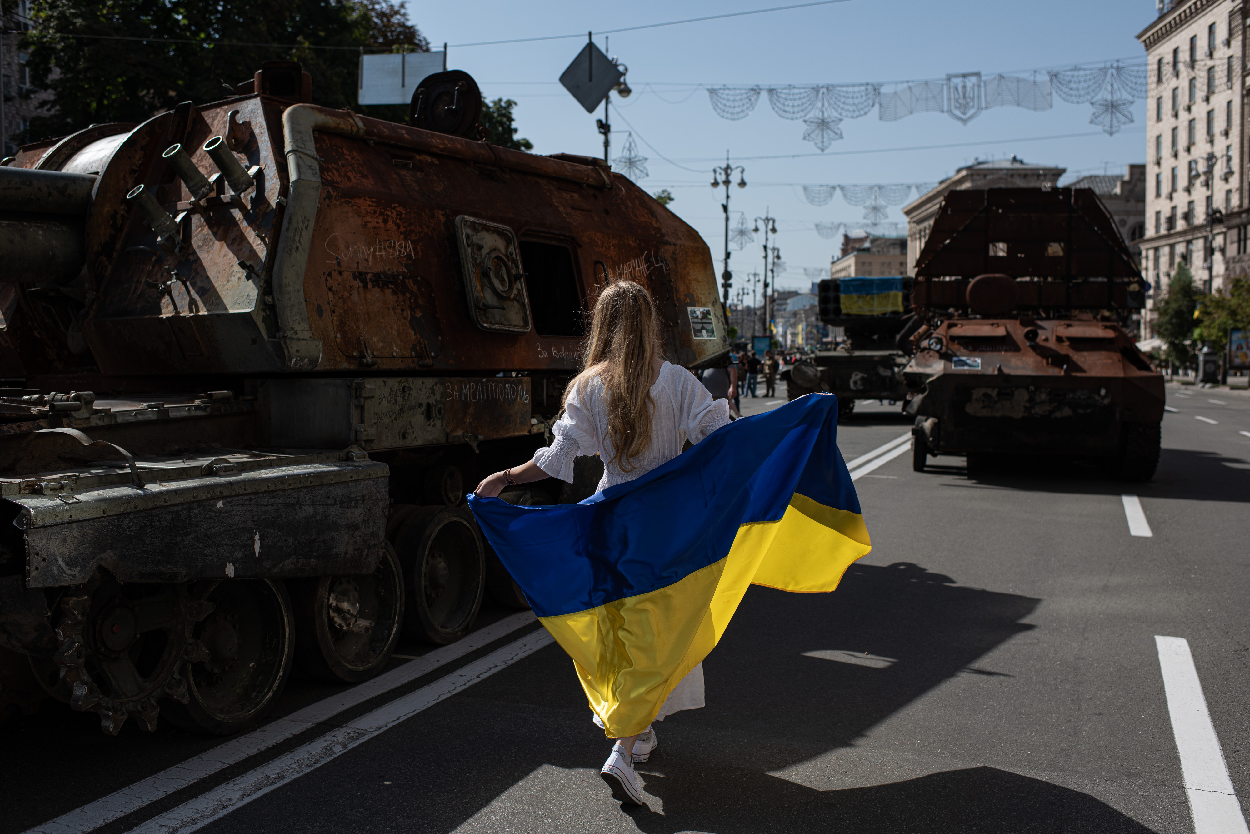 Nightly-Ukraine-082422 A photo of a woman draped in a Ukrainian flag walking by burnt Russian tanks.