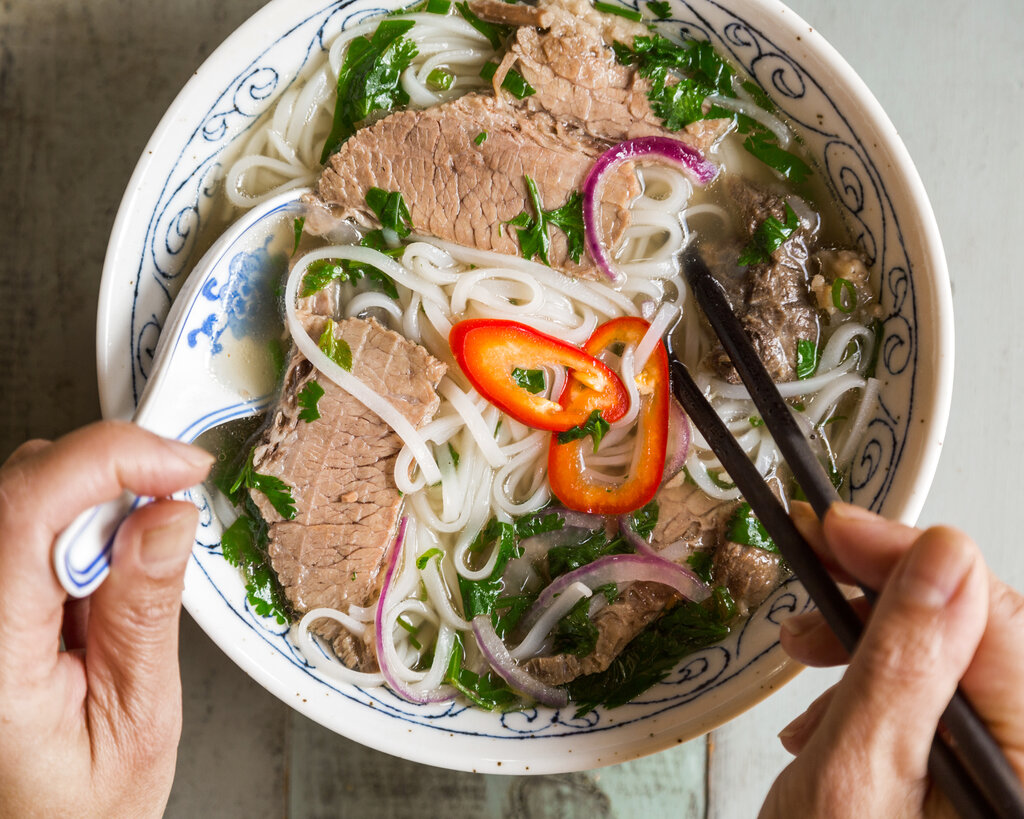 Top down view of a person's hands holding a spoon in one hand and chopsticks in the other. Both utensils are in a bowl of pho. 