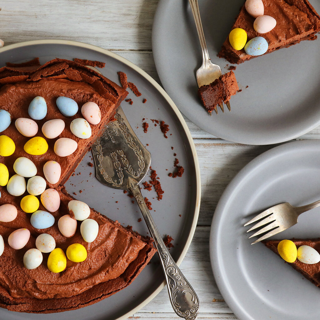 A brown cake topped with candy Easter eggs and a serving utensil sits on a gray plate alongside two other gray plates bearing slices of cake and forks.