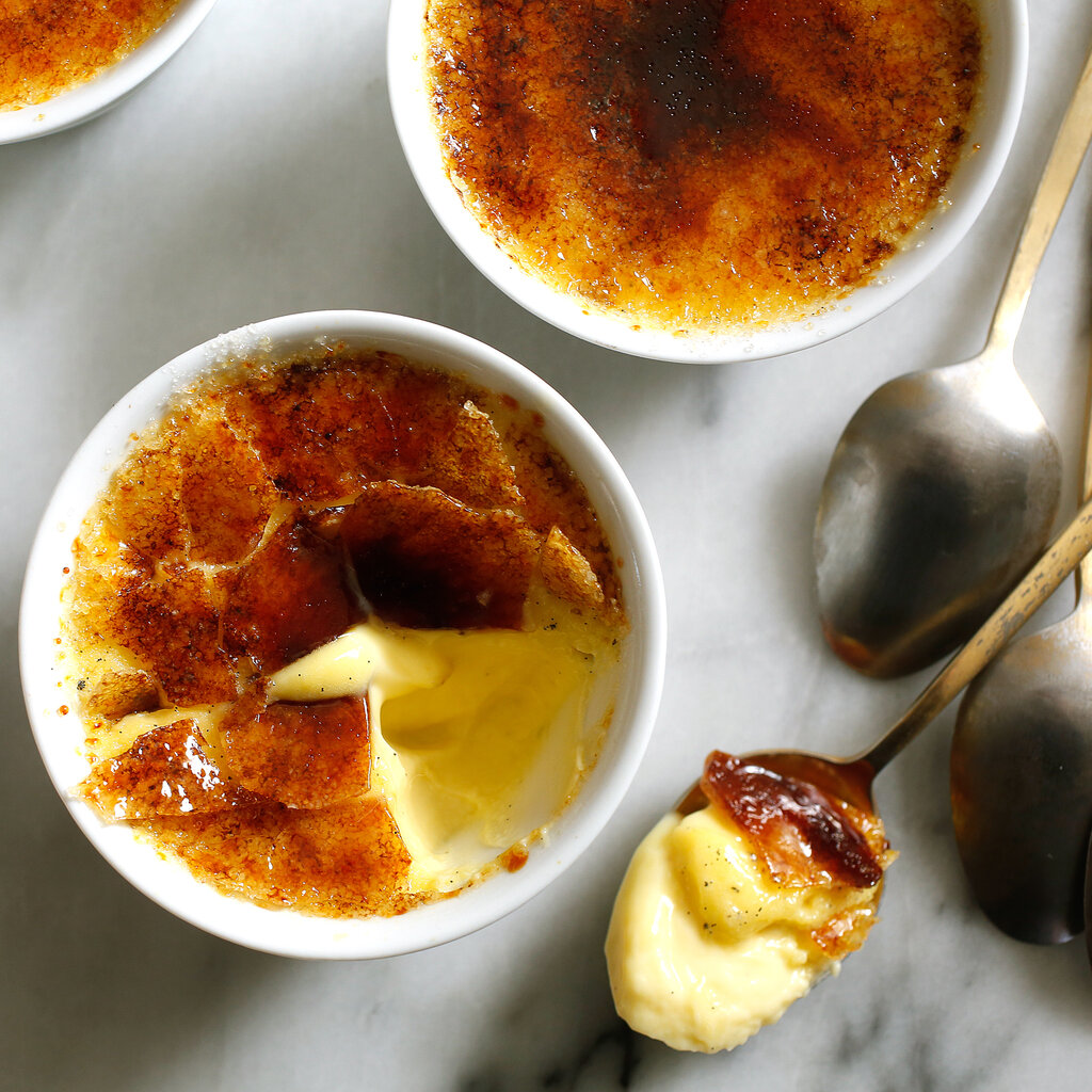 An overhead image of four ramekins filled with creme brulee. One creme brulee’s surface is cracked open with a spoonful removed. The filled spoon sits on the marble surface to the right of the dish.
