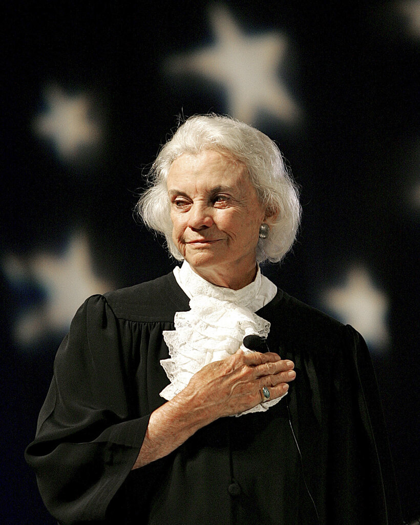 A photo of Justice O’Connor standing in sunlight in her black Supreme Court robe, her right hand over her heart. The stars of the American flag can be seen behind her, out of focus.