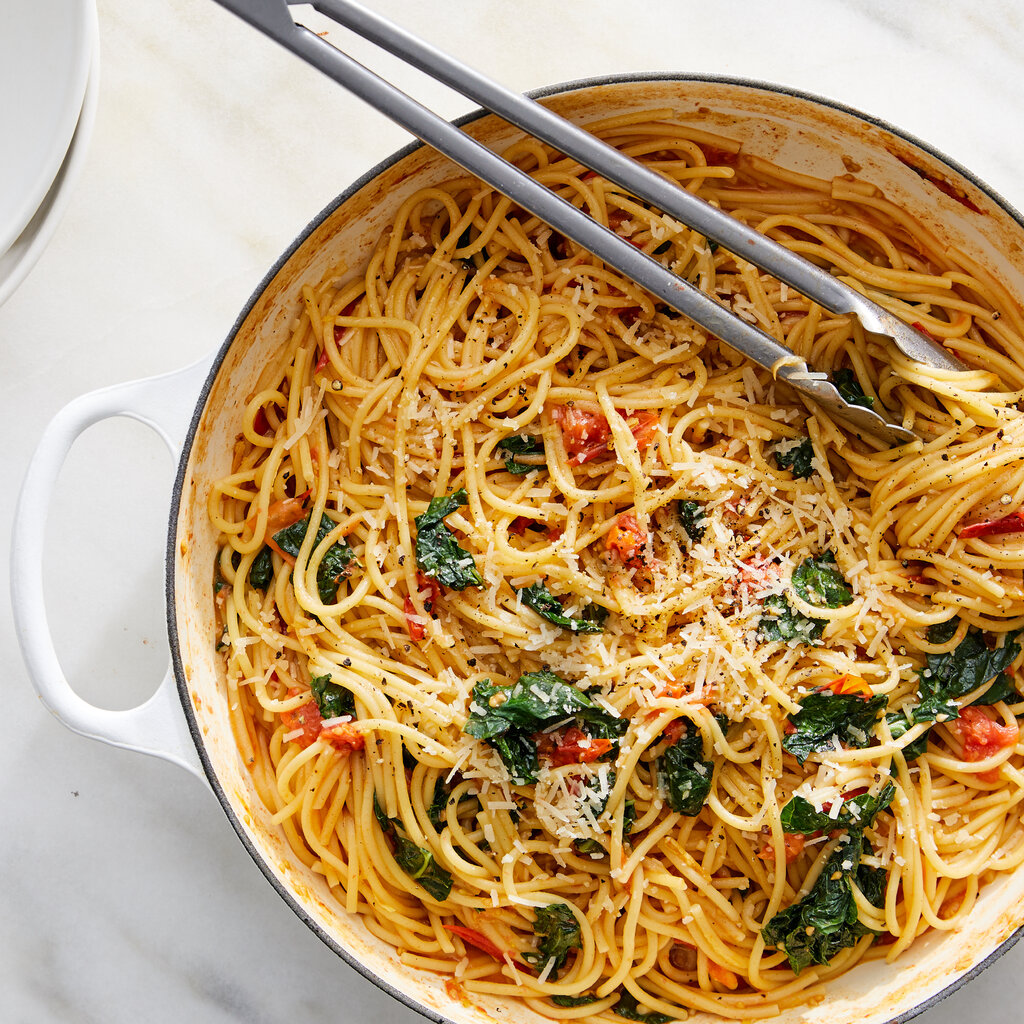 A tangle of noodles in a pot with greens and tomatoes. 