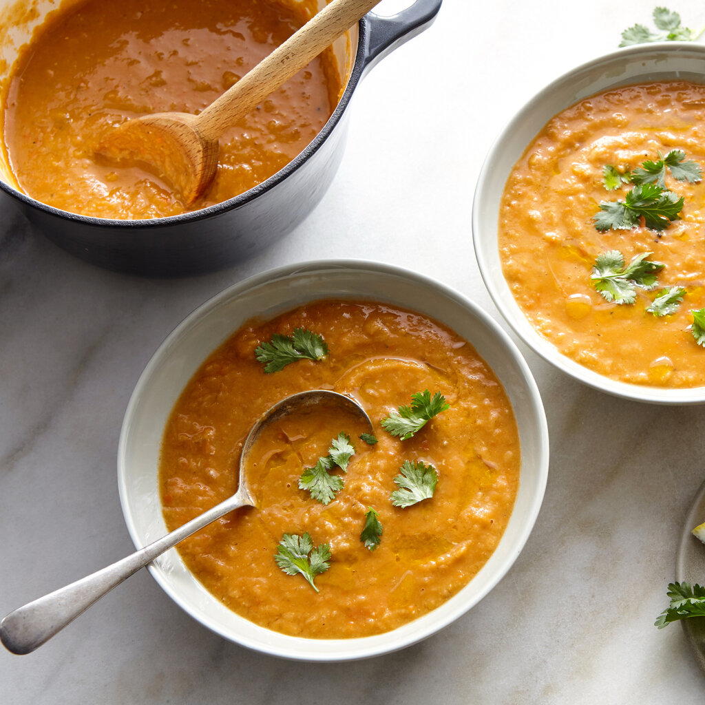 Two bowls of red lentil soup are garnished with cilantro leaves; a plate of lemon wedges and a pot of soup are nearby.