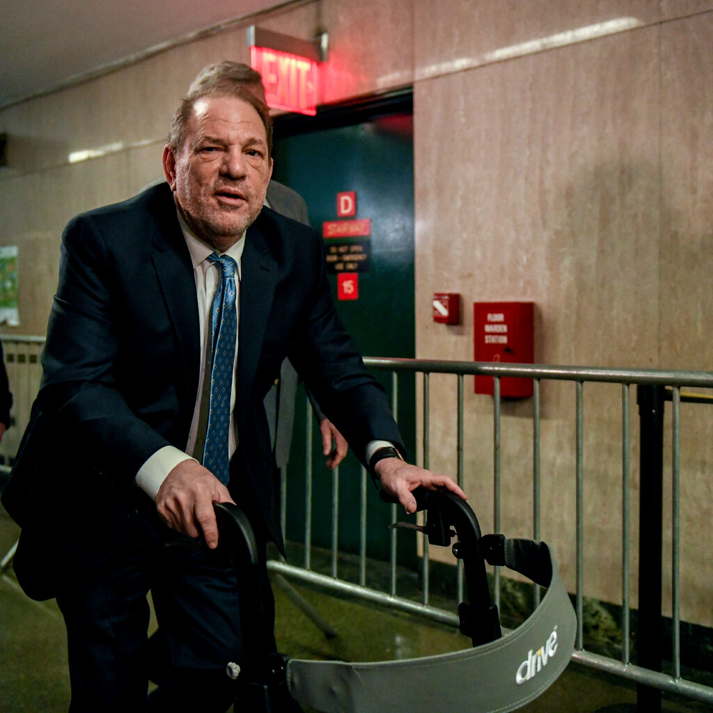 Harvey Weinstein in a suit walks down a hallway with a walker.