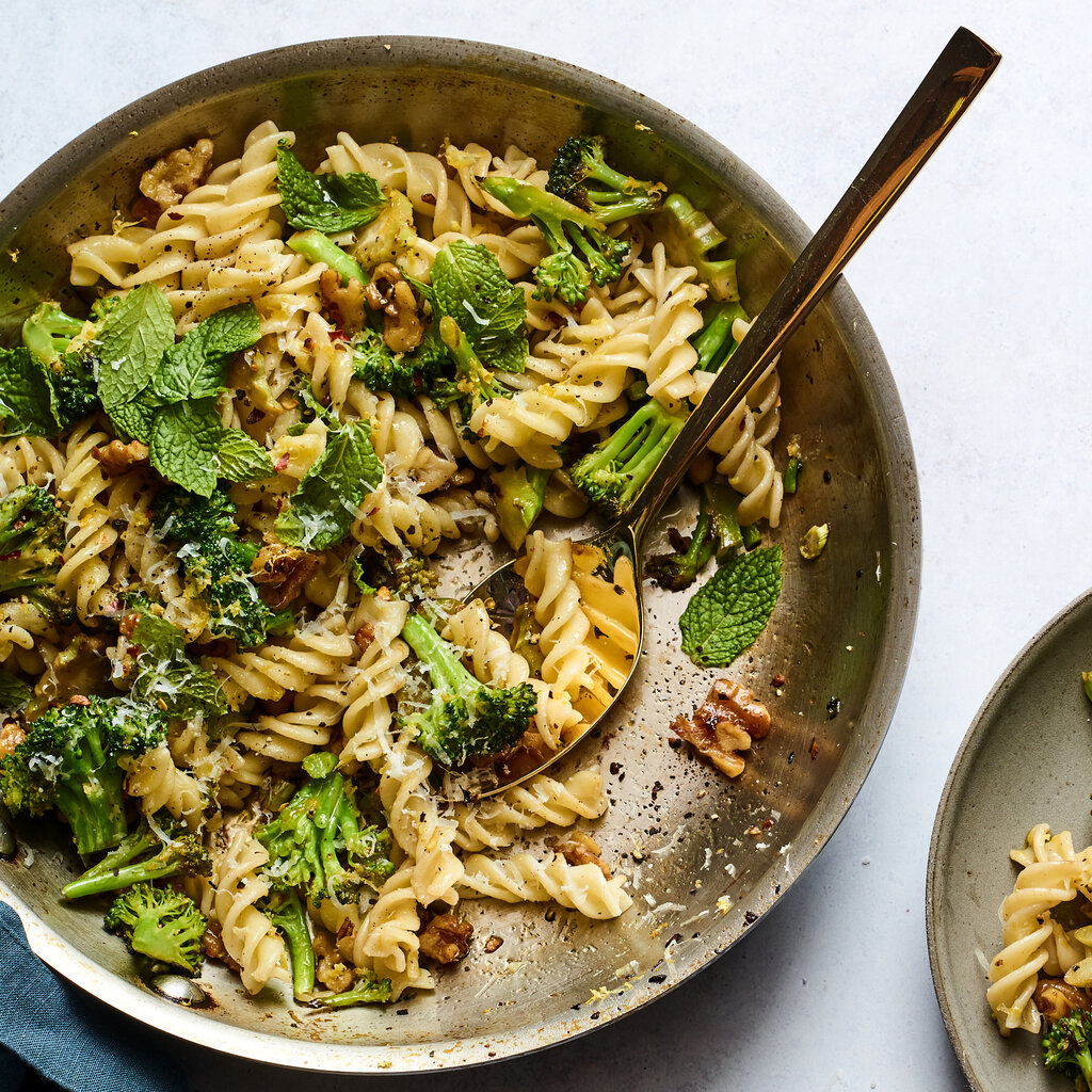 A skillet holds blistered broccoli pasta with a serving scooped out and served on a gray ceramic plate.