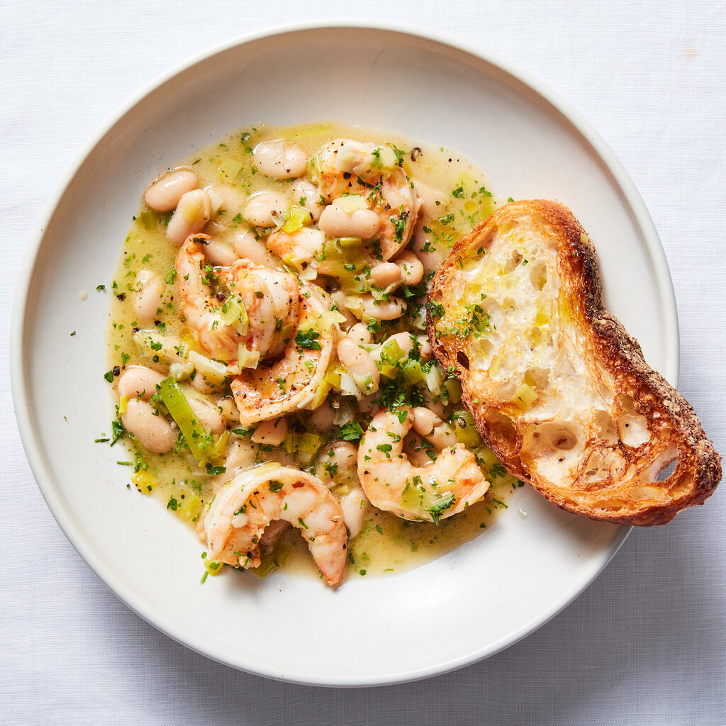 A rimmed white plate holds brothy beans and shrimp scattered with herbs and a piece of toasted bread.