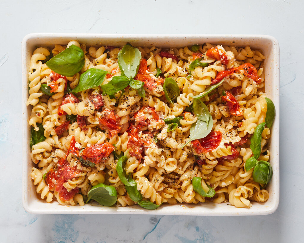 Spiral pasta, cooked cherry tomatoes, crumbled feta and basil leaves, in a rectangular white baking dish.