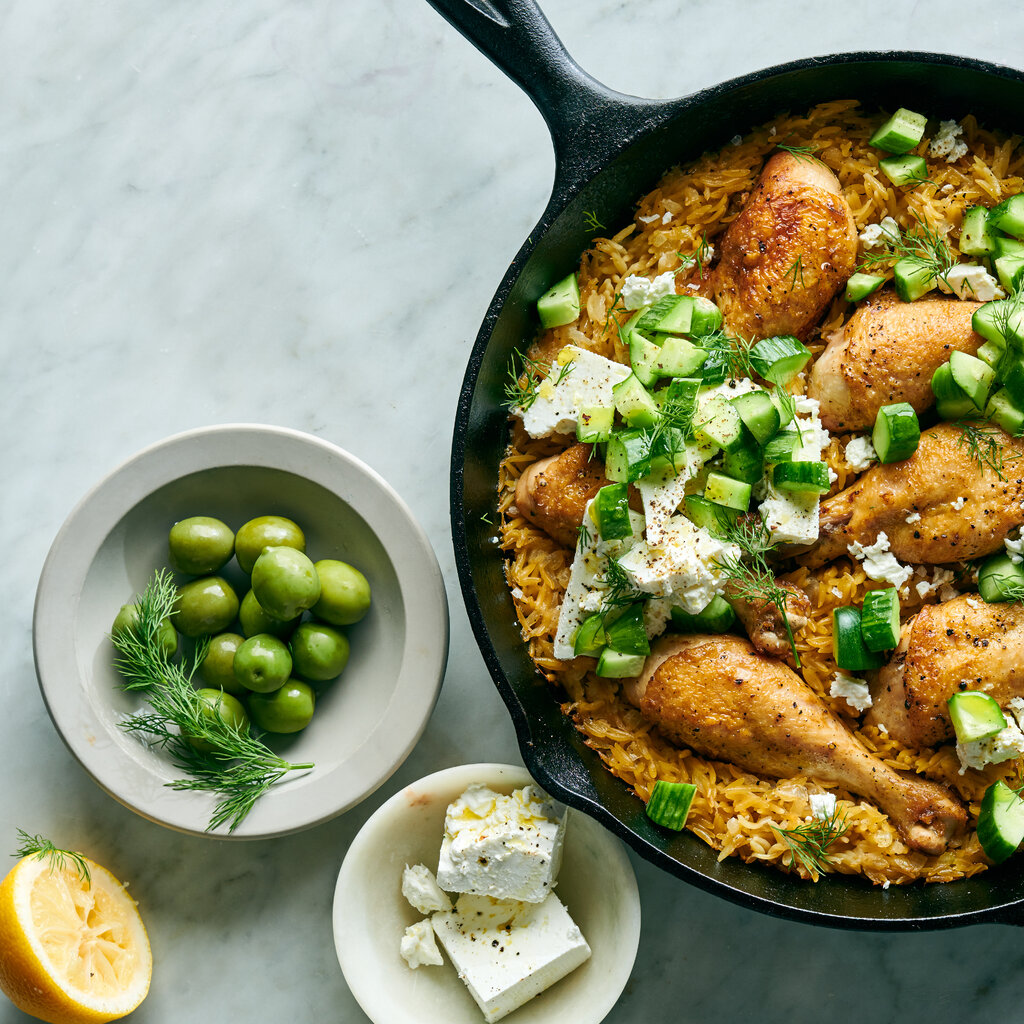 A cast-iron skillet holds chicken legs with orzo, dill and feta, with additional olives, feta and a lemon half nearby.