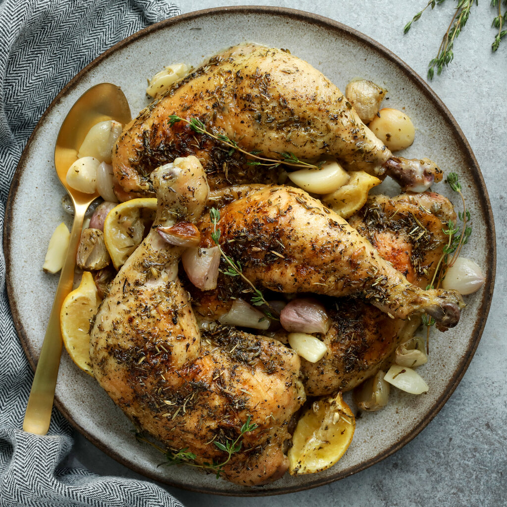 Three chicken Provençal legs are on a plate with shallots, lemon and thyme sprigs. A golden spoon is nestled in for serving.