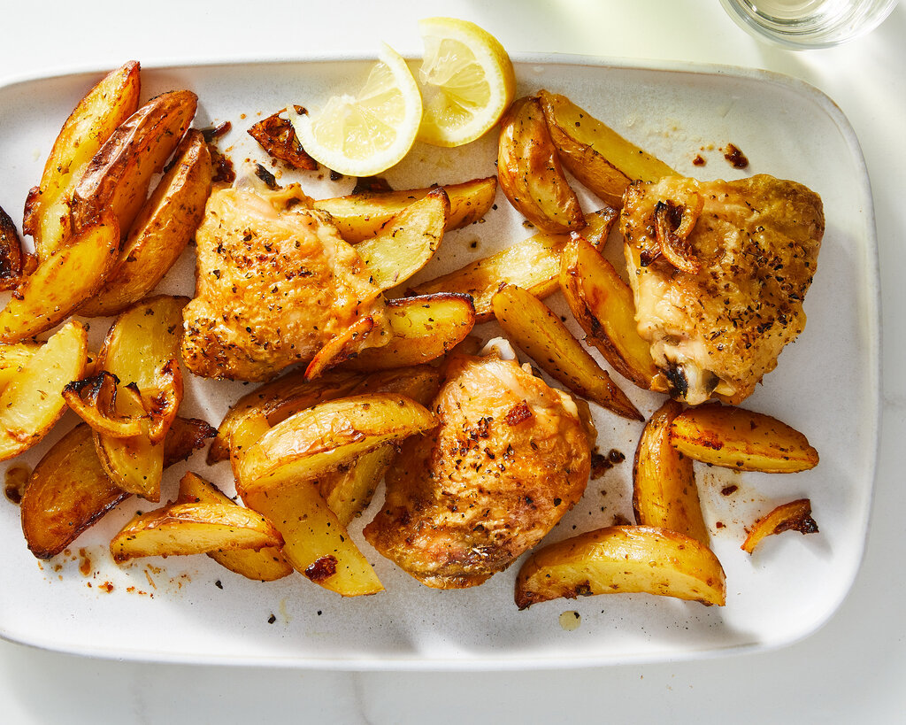 A white rectangular platter holds bone-in, skin-on chicken thighs and roasted potato wedges. Off to the top of the platter are two lemon wedges.