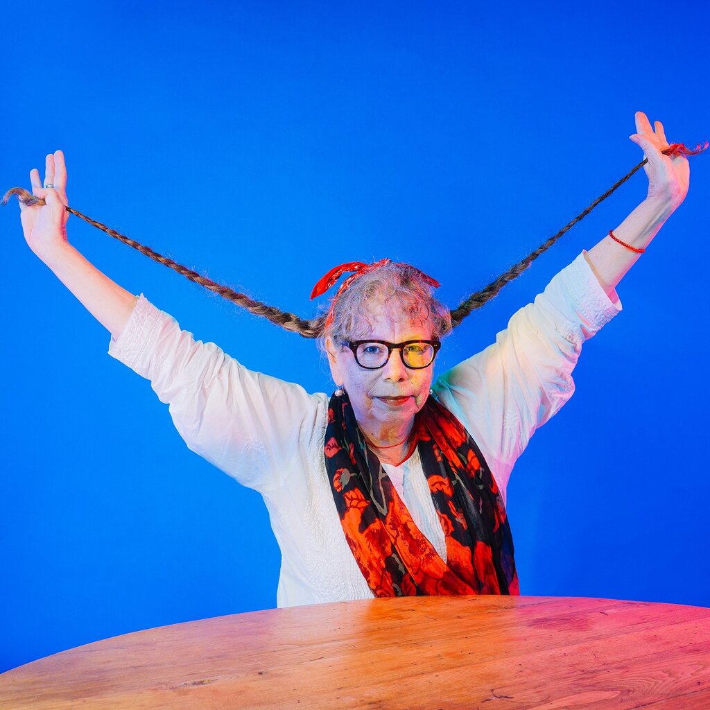 An older woman poses for a portrait against an electric blue background. Her hands are stretched out above her head, holding two braids. 