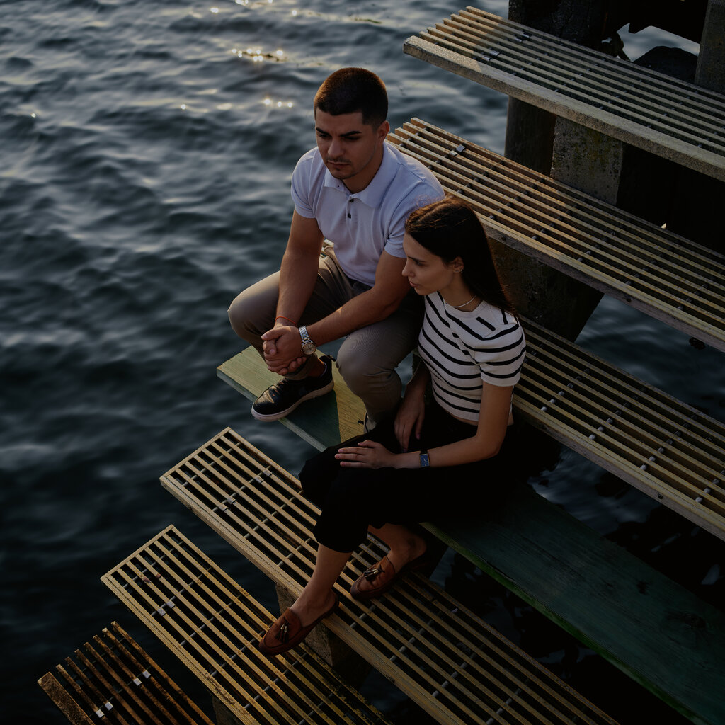 Two people sit on the steps of a staircase. Below the steps and in the background is a body of water.