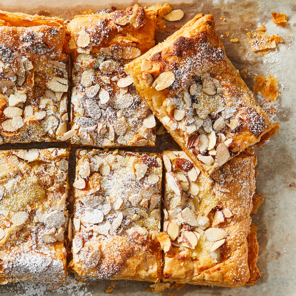 A rectangular almond croissant, showered with powdered sugar and slivered almonds, is cut into large pieces for sharing.