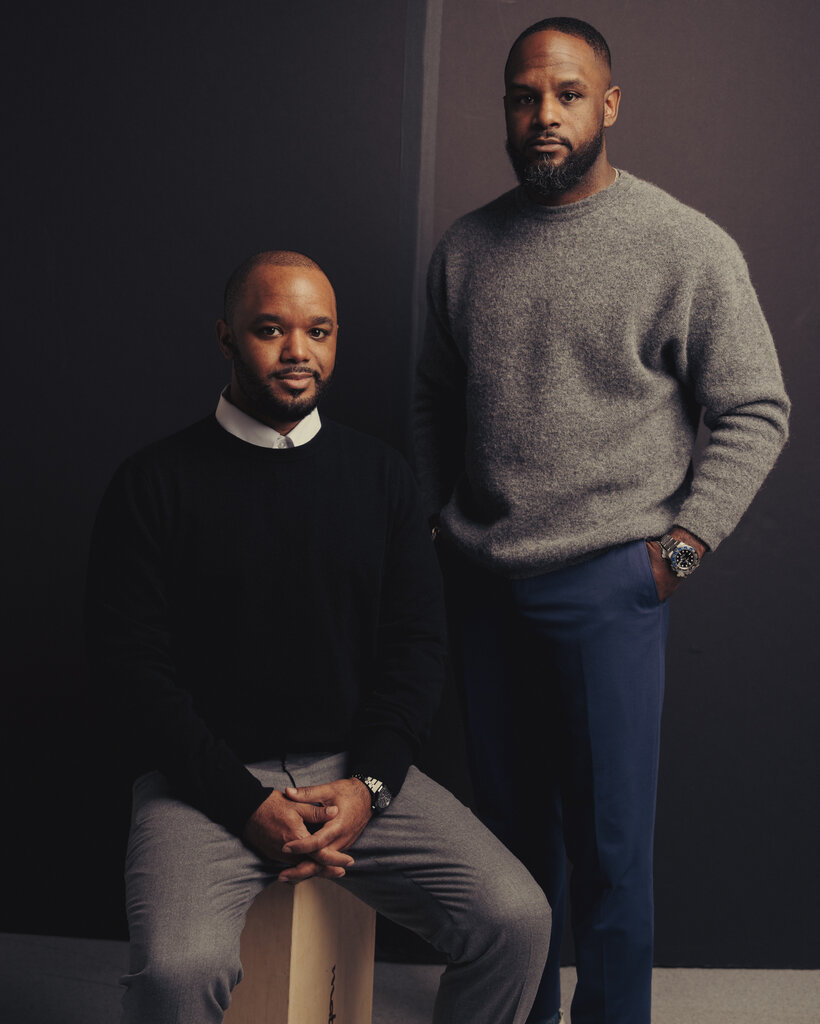 Aaron Eanes wears a black sweater and gray pants as he sits on a stool. André Eanes, wearing a gray sweater and blue pants, stands next to him.