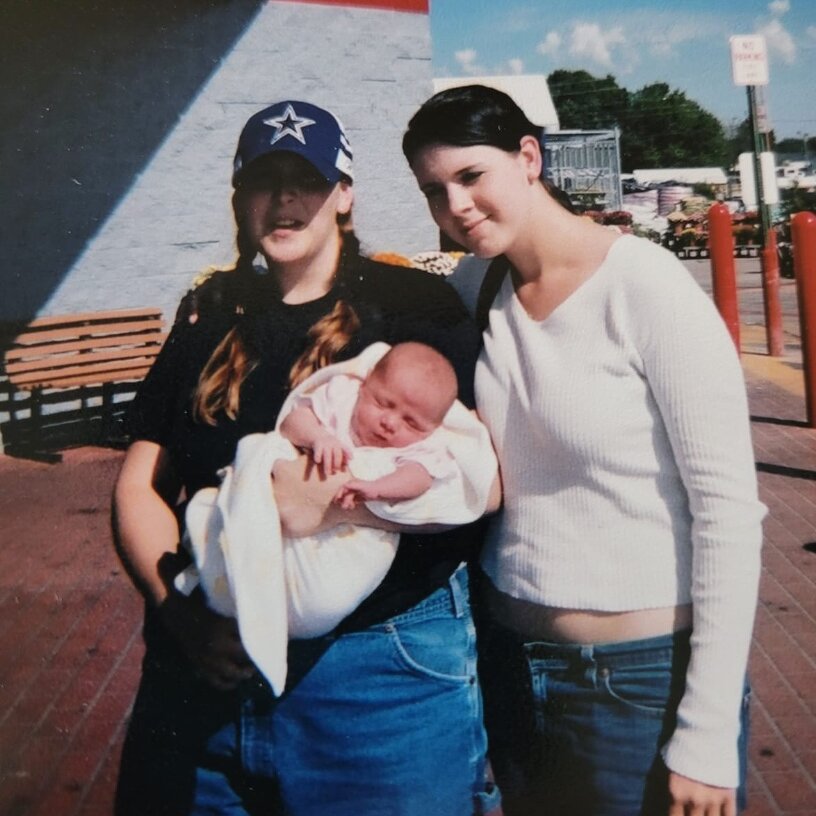 In this color photograph, two young women in blue jeans stand side by side outside near a parking lot. The woman on the left has braids and a baseball cap obscuring half her face, and she is holding an infant wrapped in a white blanket. The woman to her right has dark hair pulled back from her face and is wearing a long-sleeved white top. 