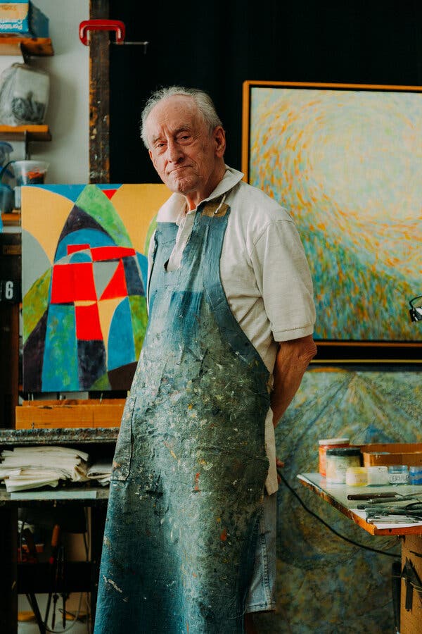 Fred Terna wearing a long blue smock with paint stains over a white shirt. His hands are behind him and he is looking at the camera. Behind him are several colorful paintings.