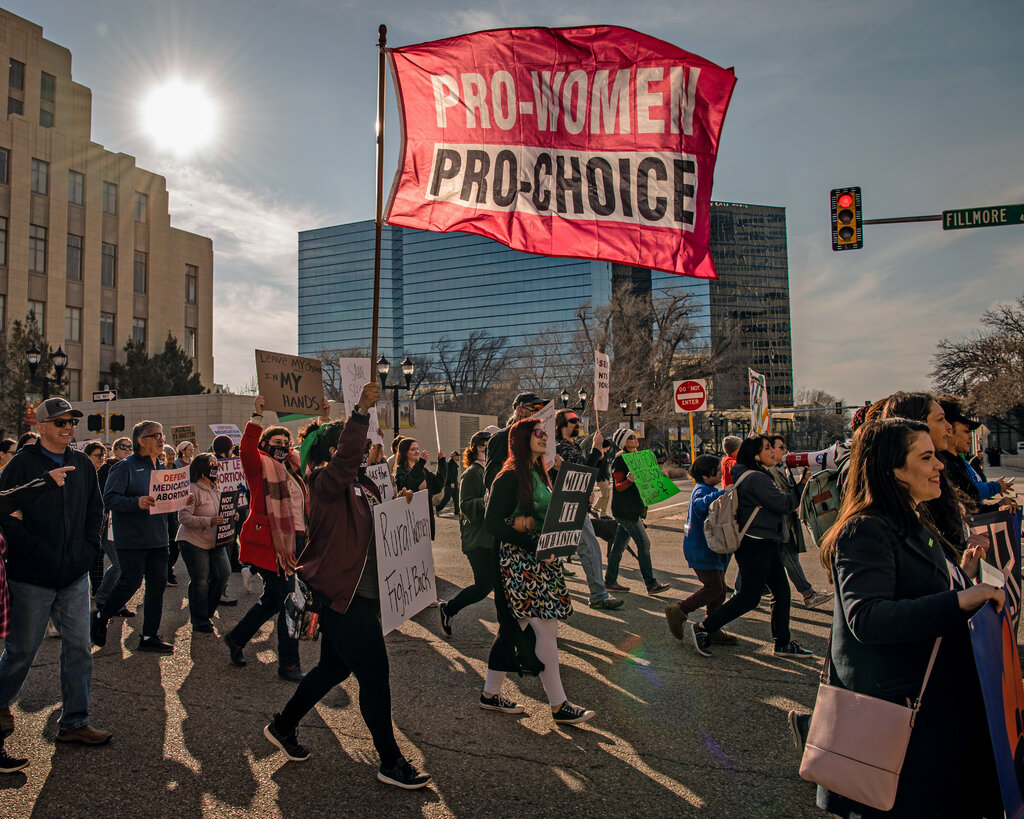 A photo shows a group of people marching through a downtown area, many of them carrying signs in support of abortion rights. One person holds a red flag that says “Pro-Women Pro-Choice.”