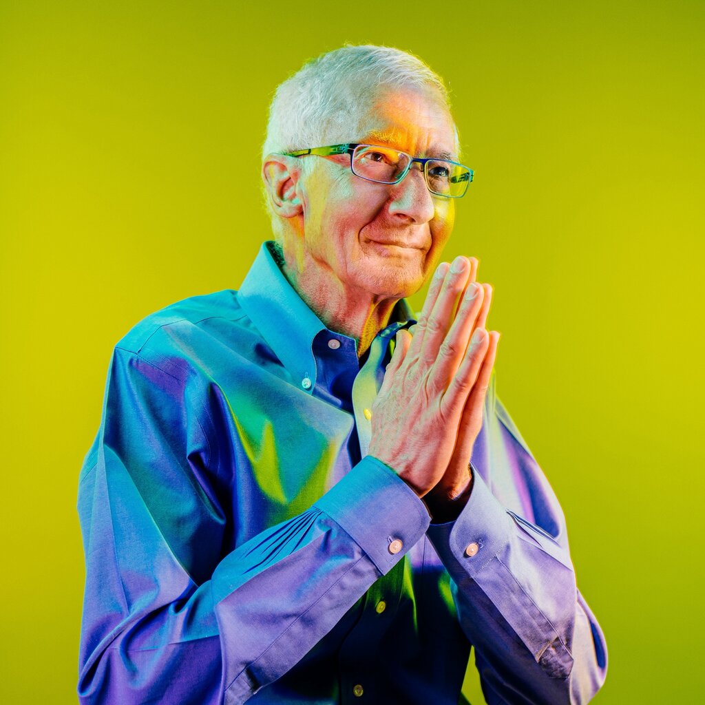 A man with gray hair and glasses, wearing a blue button down top, puts his hands together, his palms touching. 
