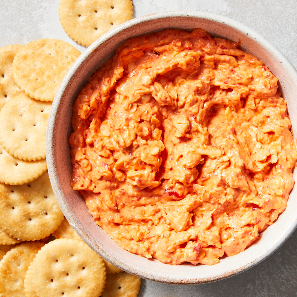 A bowl of bright orange cheese dip sits on a plate beside round crackers.