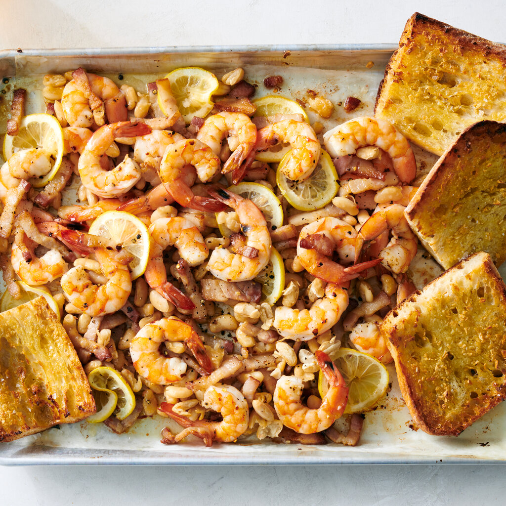 A sheet pan holds a tangle of pink roasted shrimp, white beans and lemon slices. Four toasted pieces of bread are nestled into the pan.