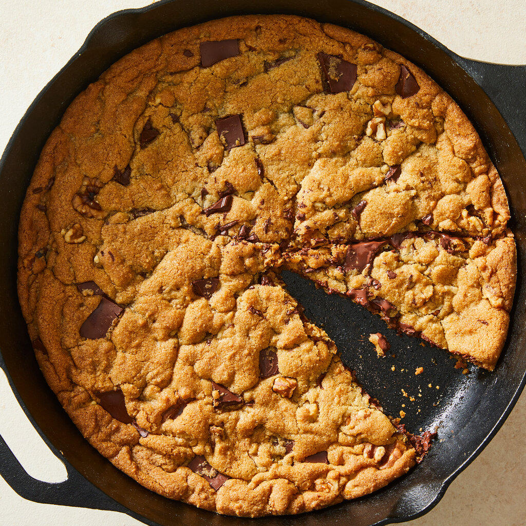 A giant skillet chocolate chip cookie has a wedge cut out of it.