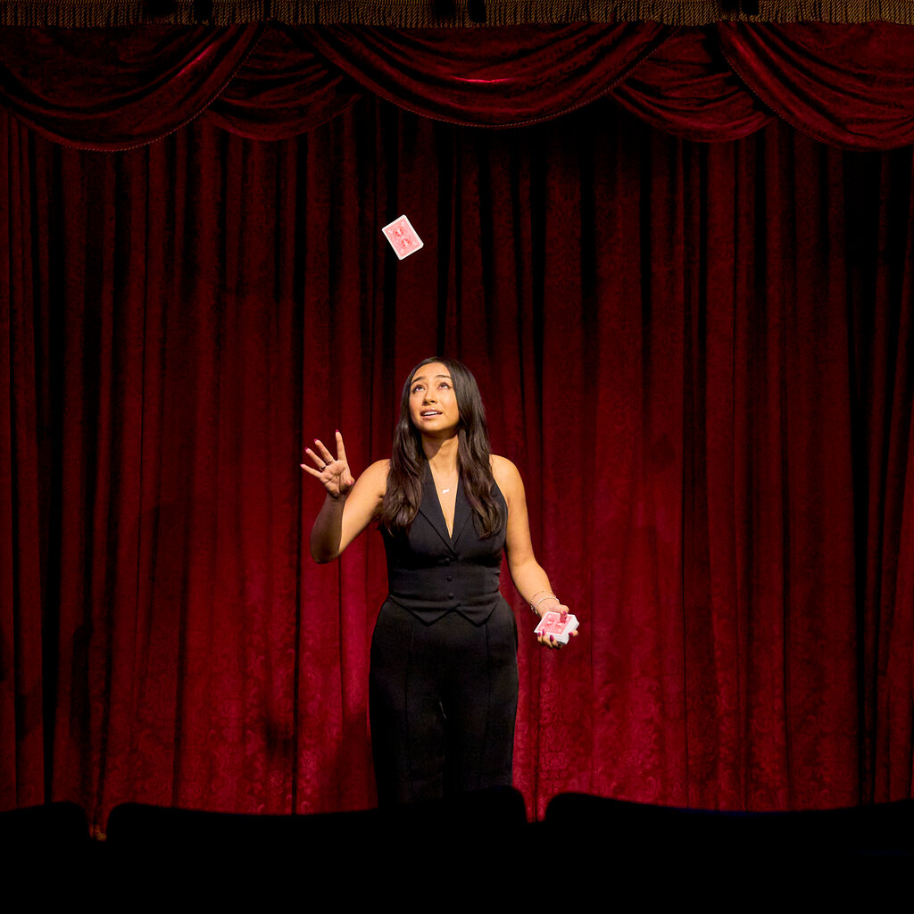 Ms. DeGuzman performing onstage in a black dress. She is holding a deck of cards in her left hand while tossing a single card into the air with her right hand.