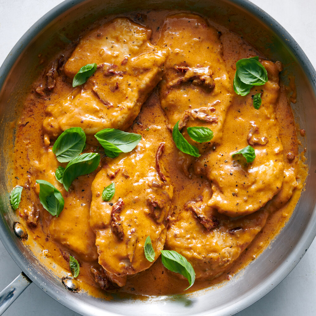 A sauté pan holds chicken breasts coated in a creamy tomato sauce and sprinkled with basil leaves.