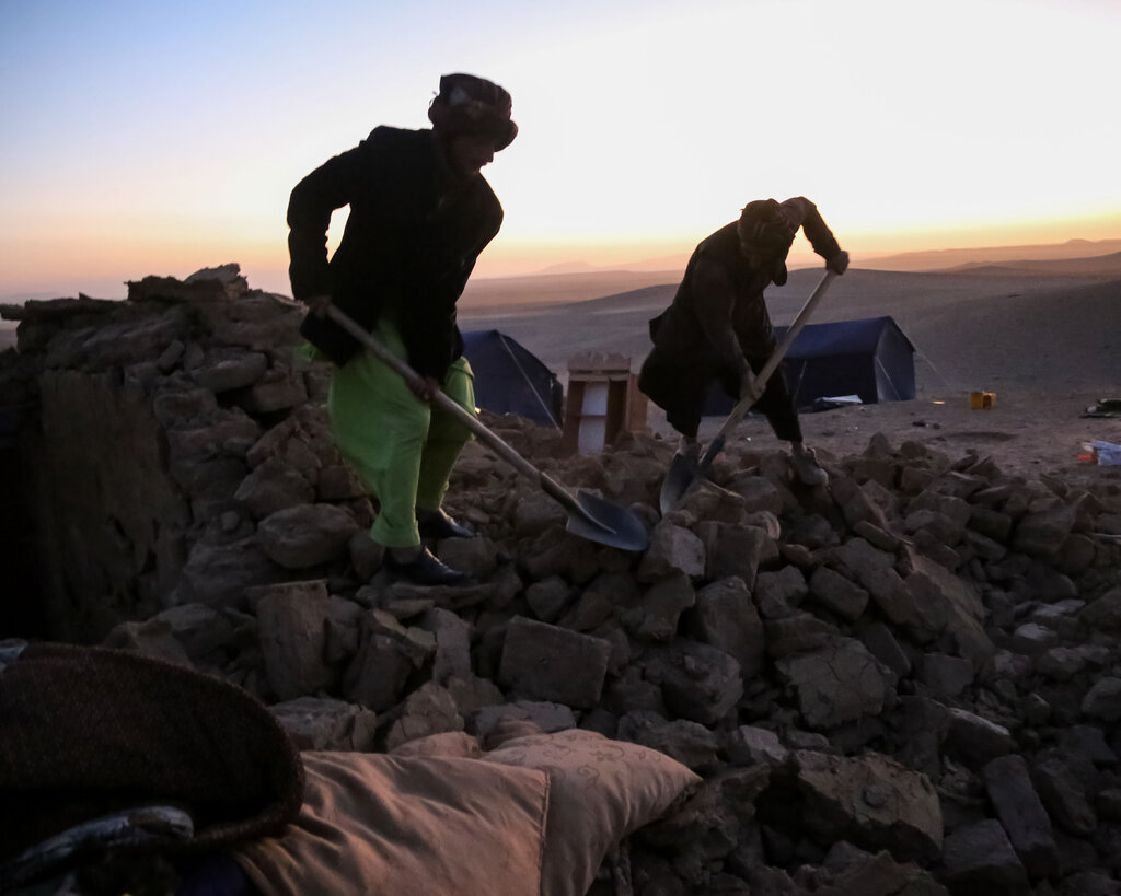 Two people use shovels to dig through rubble at dusk.