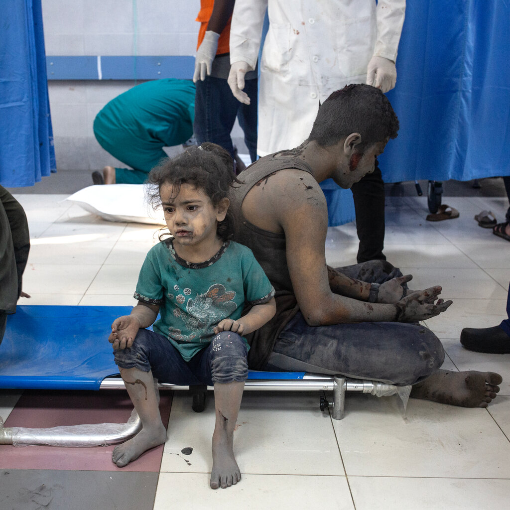 A child sits on a cot on a hospital floor, covered in dust and dirt, with bleeding wounds.