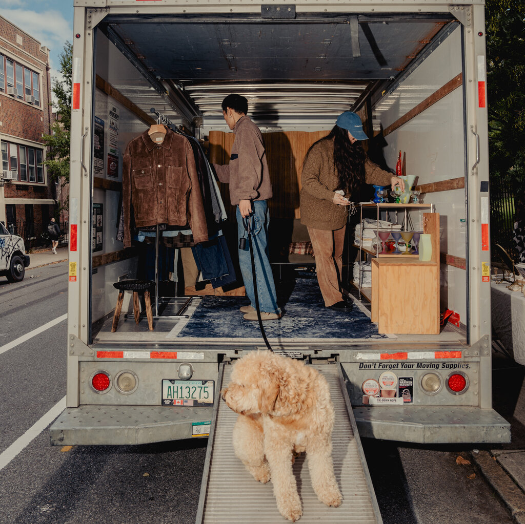 A man and a woman, both dressed in brown, look at clothing and household items in the back of a U-Haul truck. A leashed dog is sitting on a ramp leading into the truck.