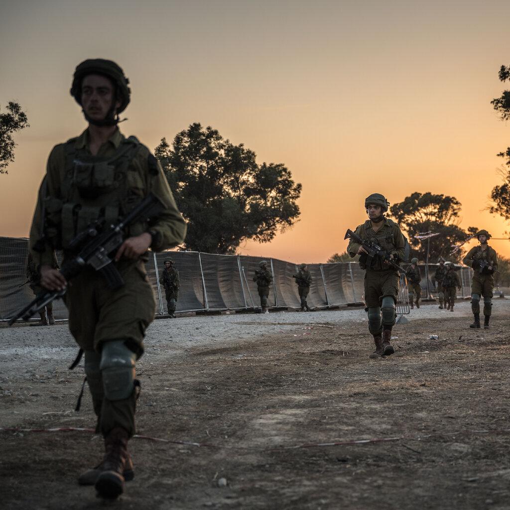 Soldiers in fatigues walk near a fence at sunset carrying guns.