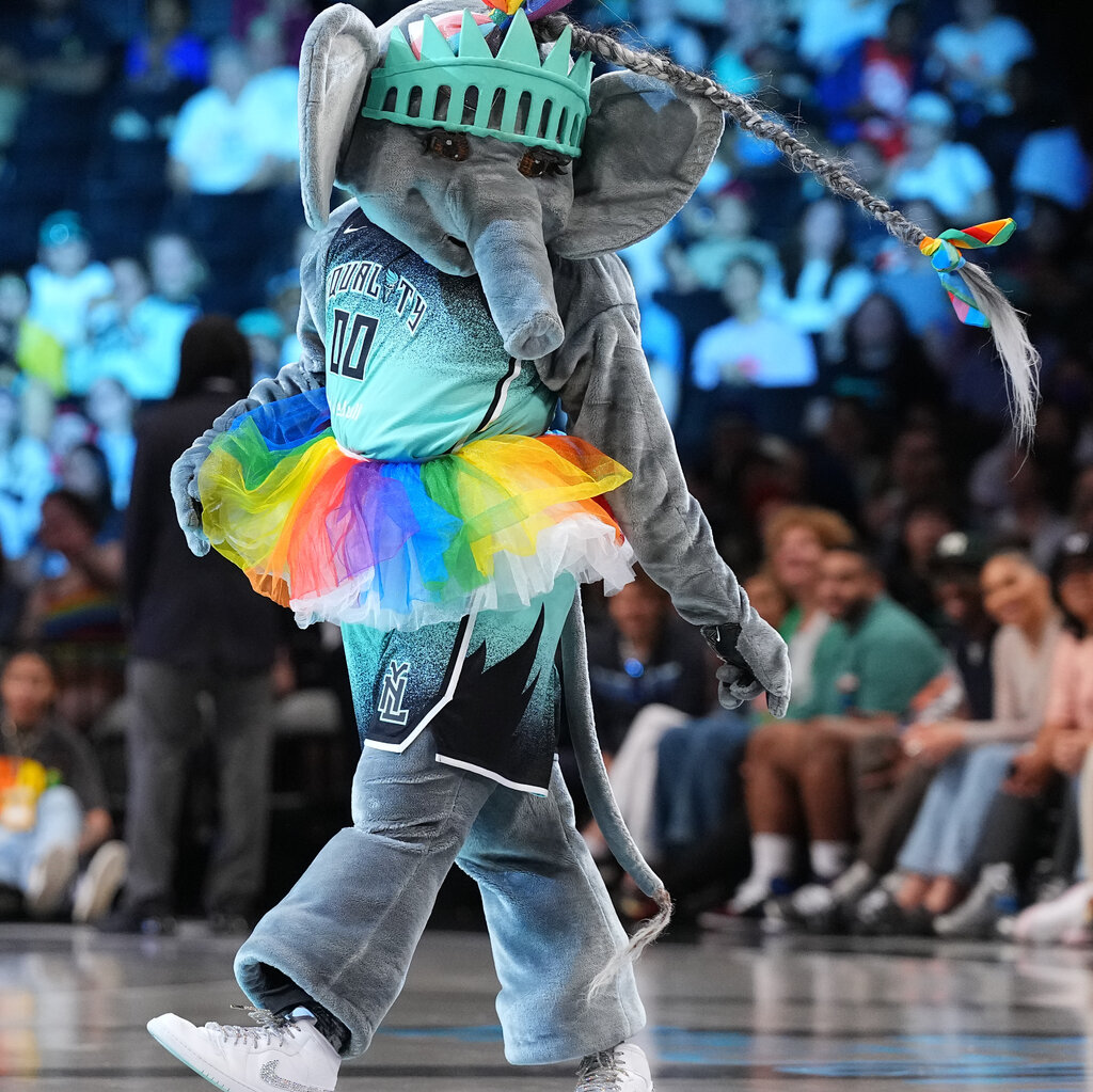 A person dressed as an elephant with a long braid adorned with rainbow-colored bows that is swinging in the air. The person is on a basketball court surrounded by seated spectators and is wearing Nike sneakers and a turquoise-and-black basketball uniform with a rainbow tutu over the elephant costume.