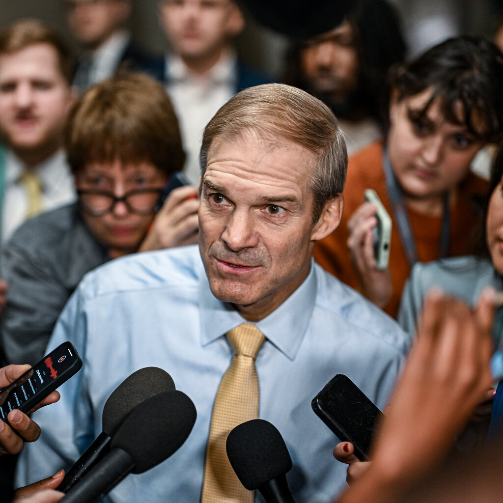 Jim Jordan in a light blue shirt and golden tie speaking into to a row of microphones and recording devices.