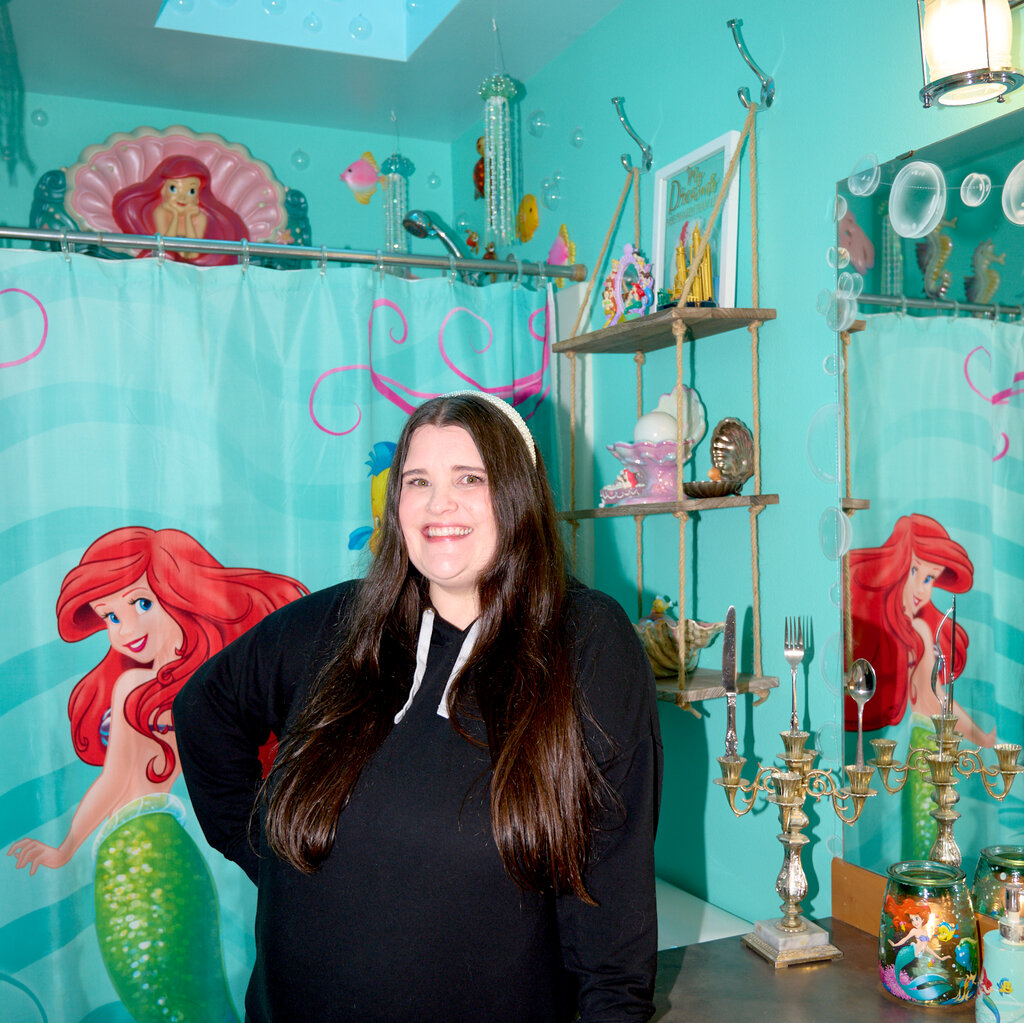 A woman with long, dark hair stands smiling in a bathroom where she is surrounded by objects that pay tribute to “The Little Mermaid.”
