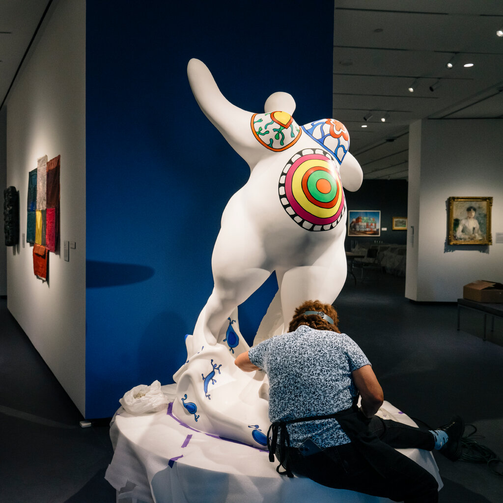 A conservator in a blue-patterned shirt and dark apron with her back to the camera cleans a large white sculpture of the pregnant Nana by Niki de Saint Phalle.