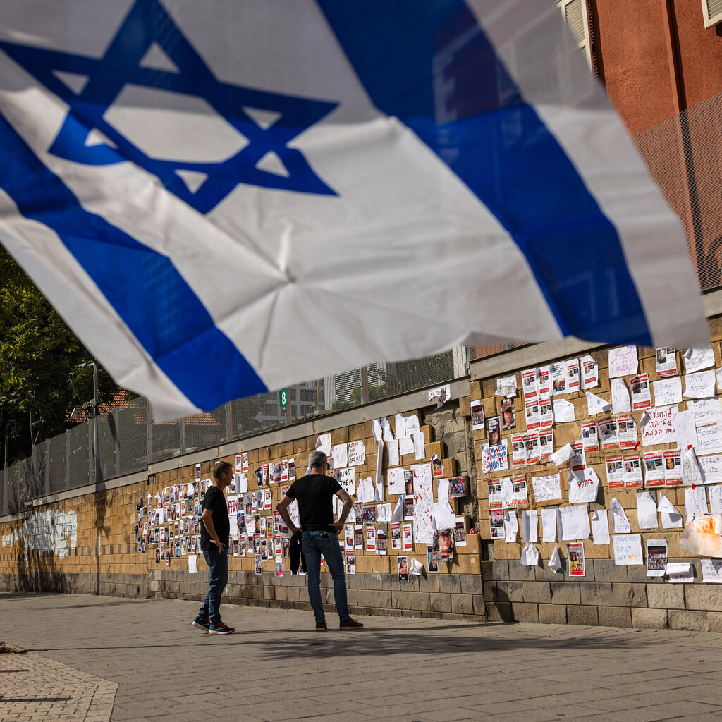 Two men stand at a wall of photographs; an Israeli flag waves in the foreground. 