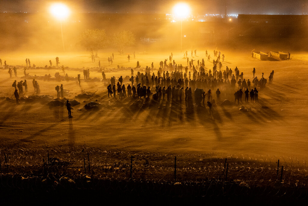 A group of people stand in a desert area, illuminated by yellow lights as the wind blows sand around them.
