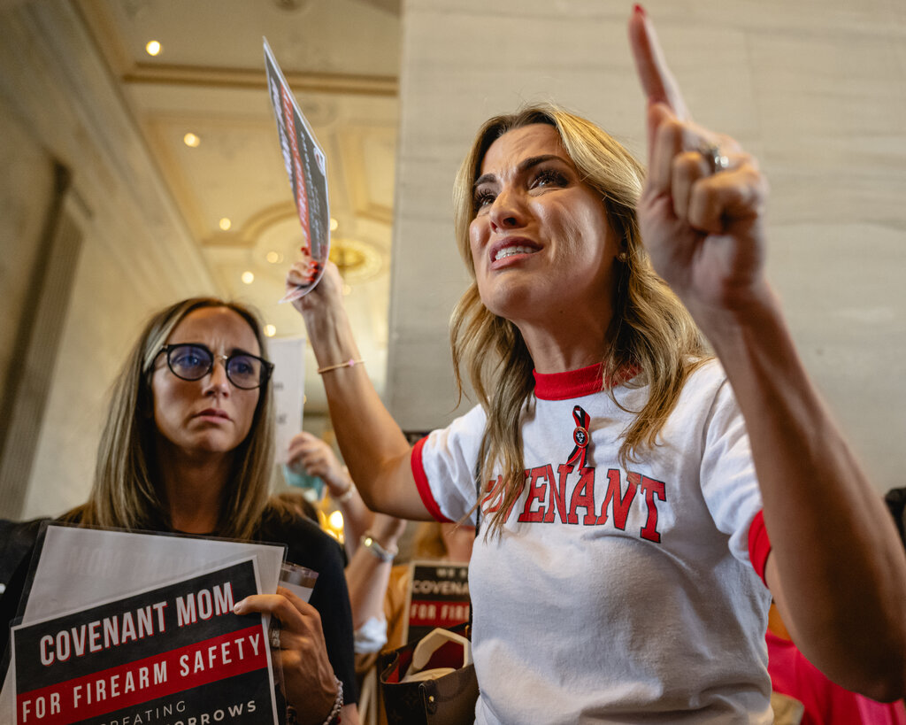 Mary Joyce raises her arms, with a look of distress on her face. She is flanked by two women holding signs that say “Covenant Mom for Firearm Safety.”