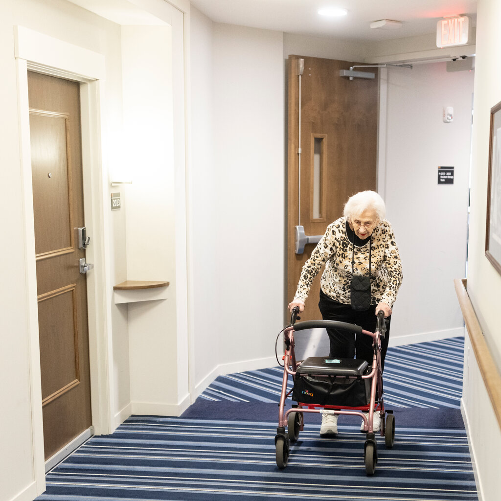 An older woman uses a rolling walker in the hallway of an assisted-living facility with white walls and three framed pictures on one side. The floor has blue striped carpeting and the doors are brown wood.
