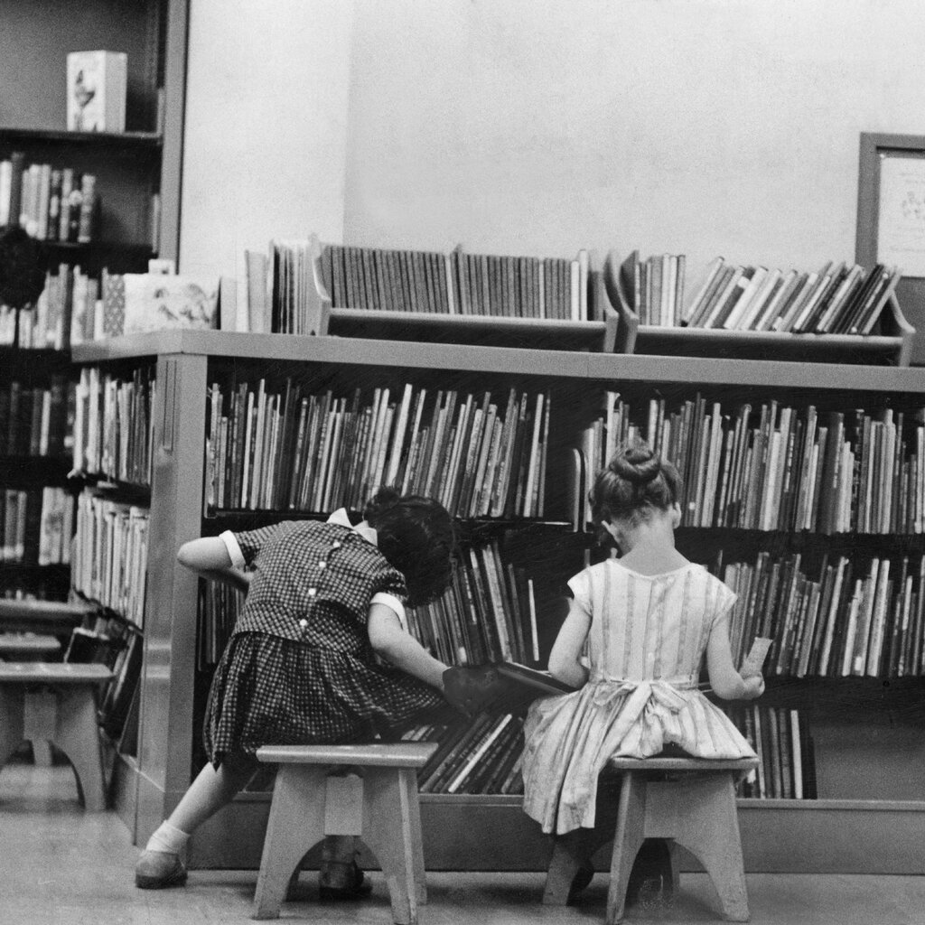 A black-and-white photo of two little girls sitting on short stools facing away from the camera toward racks of children’s books in a library.