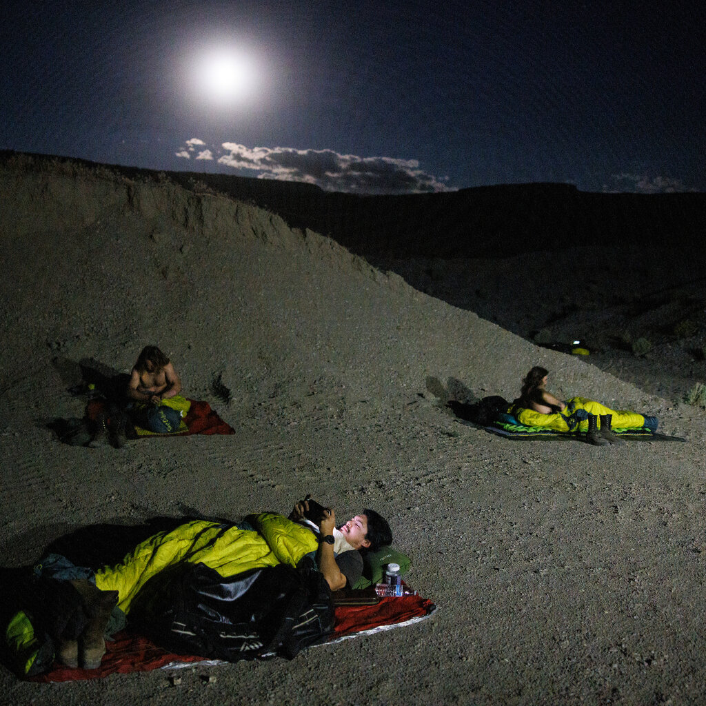 Firefighters lie in sleeping bags on dirt ground at night. The moon shines overhead. 