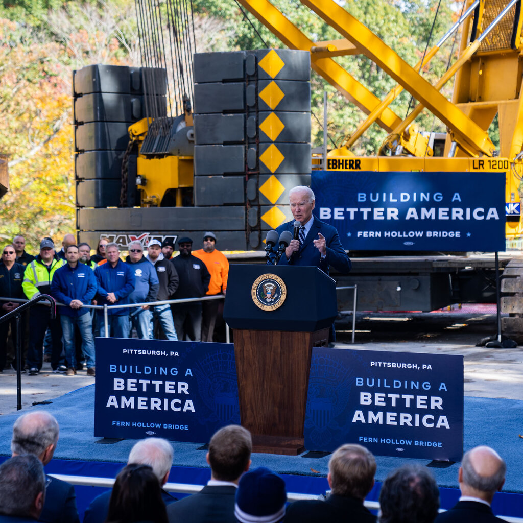 President Joe Biden giving a speech at a podium in front of workers and a large yellow construction crane.