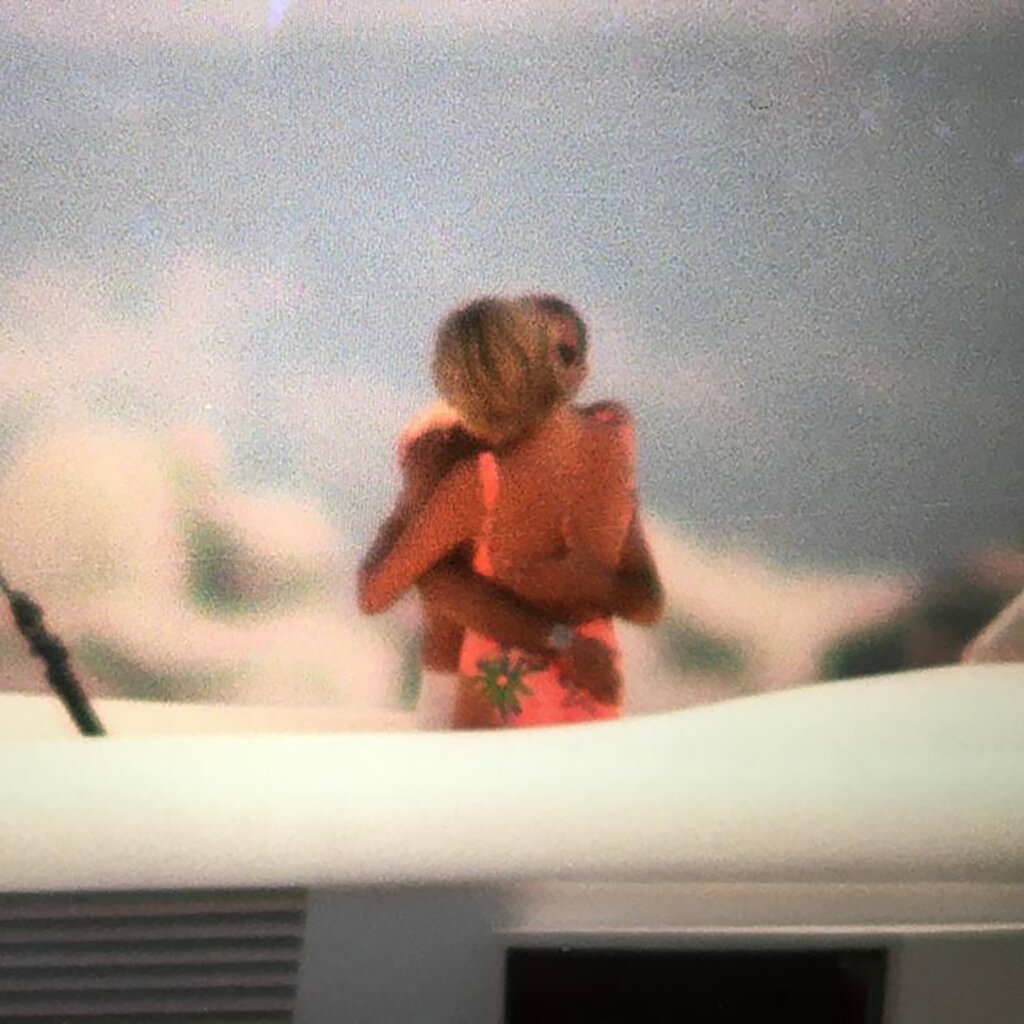 A blurry photo of Princess Diana in a pink swimsuit embracing Dodi Fayed, who is shirtless and wearing sunglasses, on a yacht.