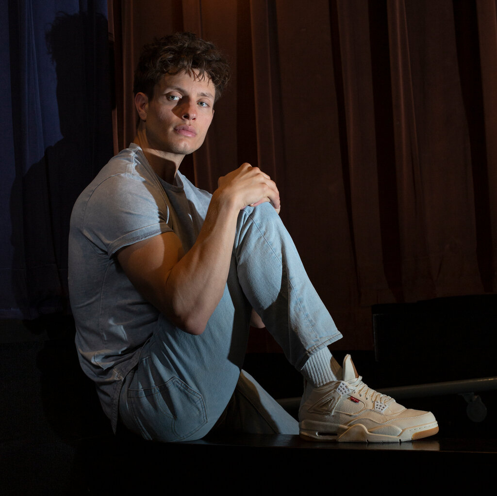 A man with brown hair, wearing a gray shirt, light blue jeans and white sneakers, sits on a stage with a spotlight on him. 