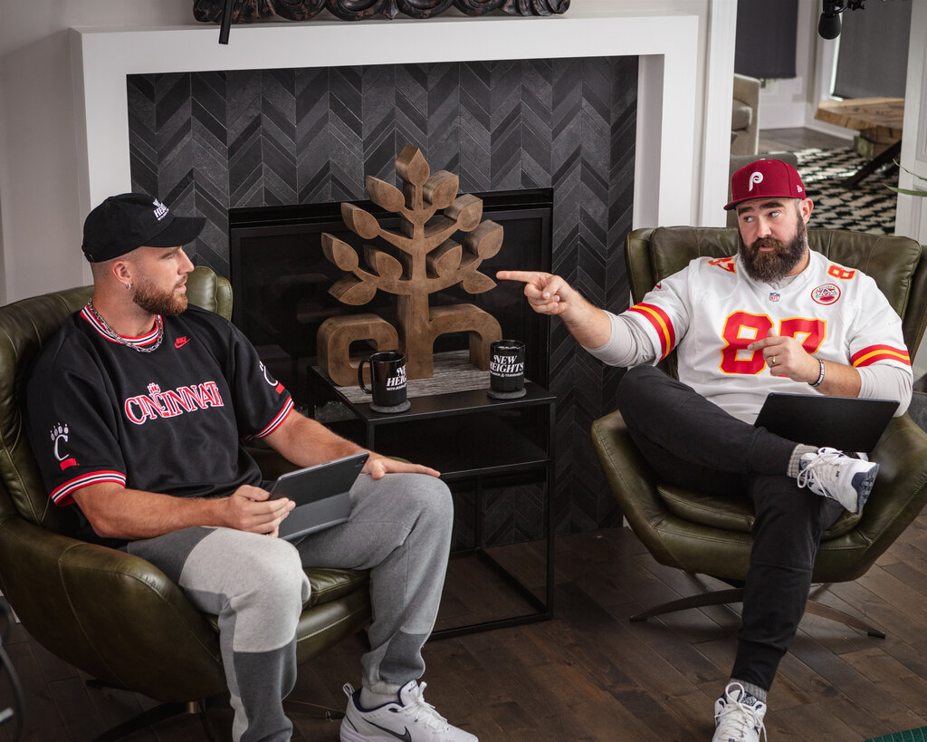 Travis Kelce (left) and his brother Jason, both wearing sports jerseys, sit in chairs with coffee cups between them. Jason is pointing at Travis.