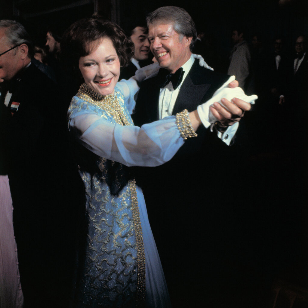 Rosalynn Cater in a blue dress and Jimmy Carter in a suit holding hands and dancing together.