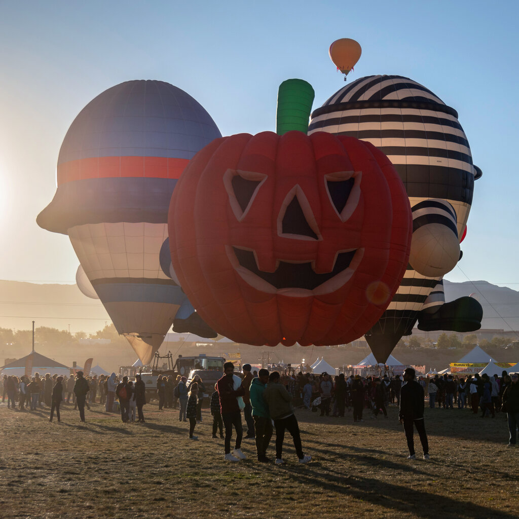 An orange hot air balloon shaped like a grinning jack-o’-lantern with two other whimsical balloons, one with blue, red and white stripes and the other with black and white stripes, in the early-morning light.