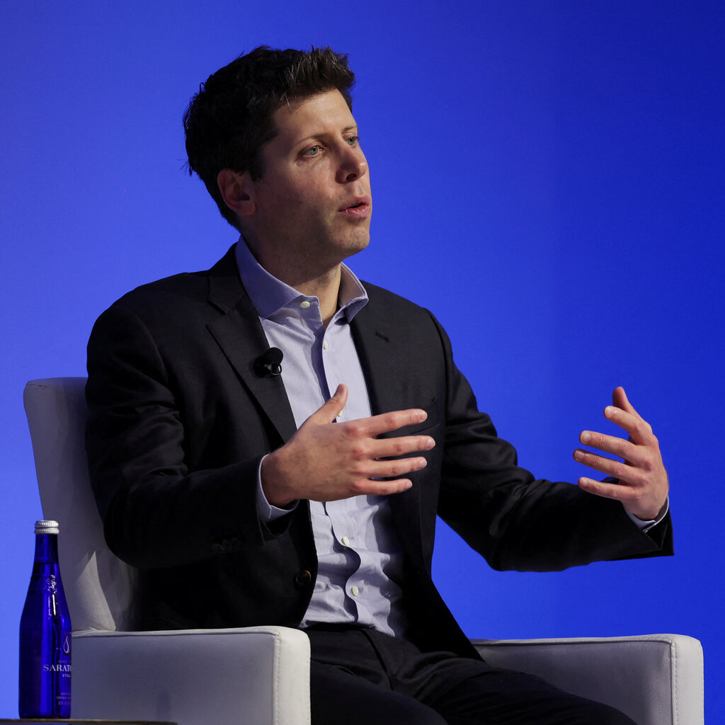 Sam Altman, in a suit and lilac shirt, sits in front of a blue background. 