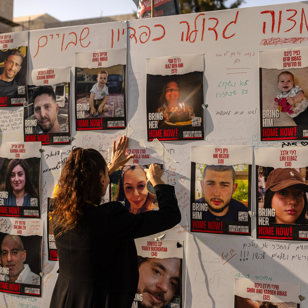 A person hanging up a flier on a wall covered with photos of hostages. The text on the photos says “Bring him home now!” and “Bring her home now!”