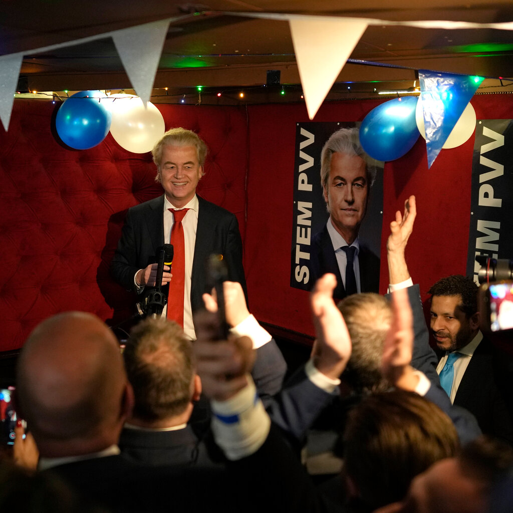 Geert Wilders, in a dark suit and red tie, stands in a small room holding a microphone as people in front of him cheer. Balloons and streamers are overhead.